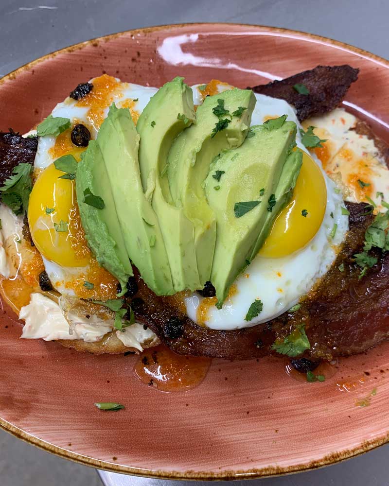 an everything bagel topped with bacon, eggs and avocado
				