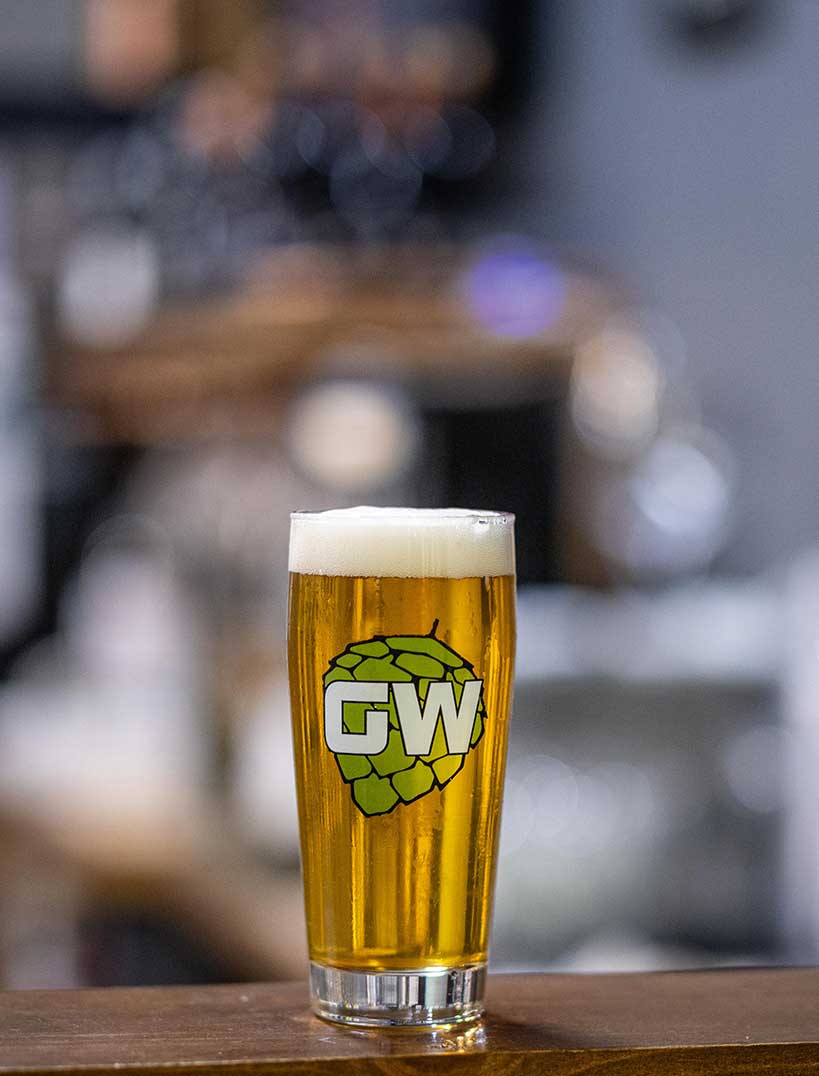 a pint of beer with a GW label on the glass in front of a blurry background 
				