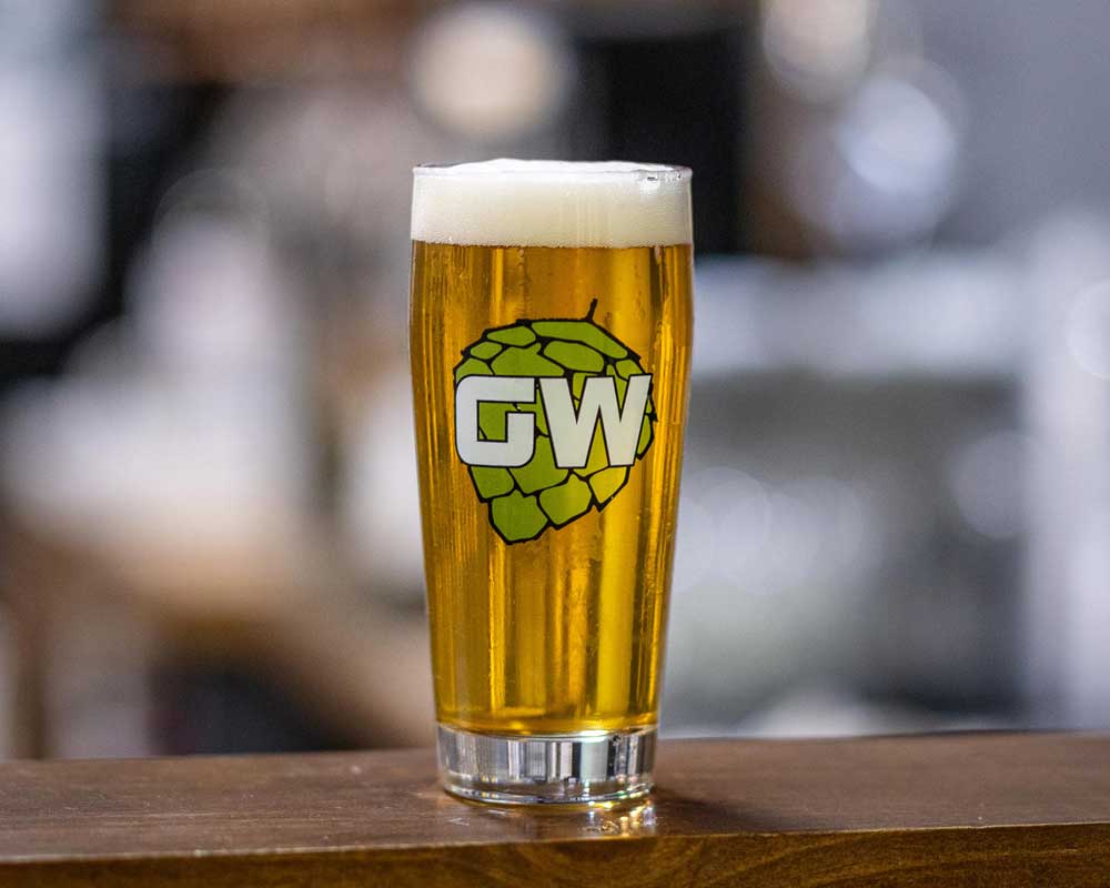 a pint of beer with a GW label on the glass in front of a blurry background 
			